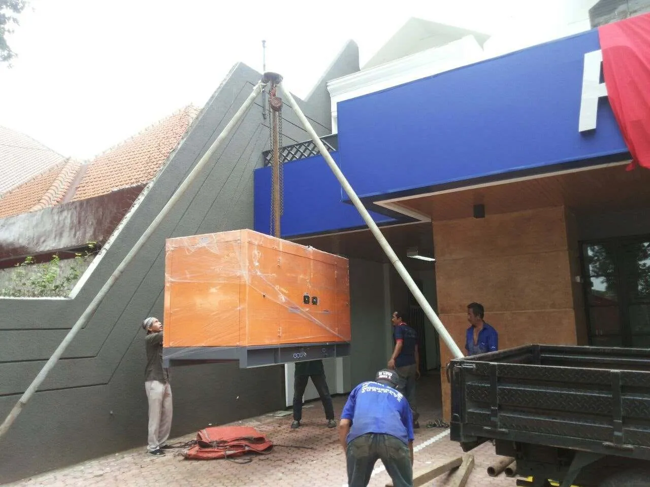 Genset Yanmar terbungkus plastik diangkat menggunakan sistem katrol di area bangunan komersial